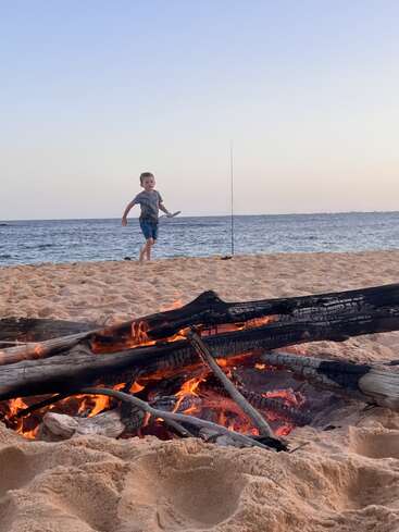 Un jeune garçon court sur une plage de sable au coucher du soleil, près d'une canne à pêche, avec un feu de camp de bûches brûlantes brillant chaleureusement au premier plan. En arrière-plan, l'océan.