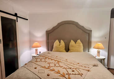Cette chambre à coucher confortable est dotée d'une grande tête de lit rembourrée, d'une literie beige aux détails texturés, de deux oreillers jaunes, de tables de chevet assorties et de lampes chaudes et lumineuses.