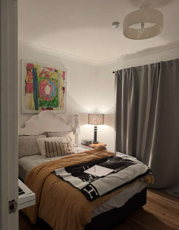 Une chambre à coucher confortable avec un lit bien fait, une couverture moutarde, des oreillers, une lampe de chevet, un tableau abstrait au-dessus de la tête de lit, des rideaux gris et un clavier de piano.