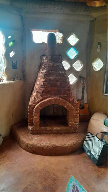 Esta acogedora habitación cuenta con una chimenea rústica de barro, ventanas de cristal de colores, una guitarra acústica, una iluminación tenue, detalles de mosaico en el suelo y una bolsa de tela a cuadros en el suelo.