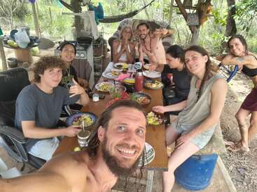 Un grupo de amigos disfruta de una comida al aire libre alrededor de una mesa rústica de madera, rodeados de árboles. Todos parecen felices, relajados y enfrascados en la conversación y las risas.