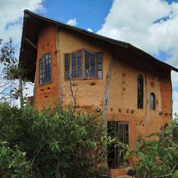 Esta imagen muestra una casa rústica de adobe de dos plantas con ventanas de madera, rodeada de exuberante vegetación bajo un cielo azul intenso con algunas nubes dispersas.
