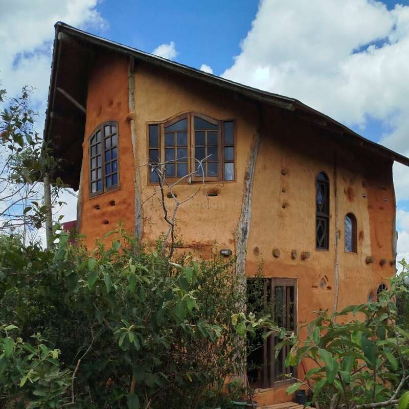 Esta imagen muestra una casa rústica de adobe de dos plantas con ventanas de madera, rodeada de exuberante vegetación bajo un cielo azul intenso con algunas nubes dispersas.