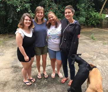 The image shows four women standing together with two dogs in front of them, posing for a photo in a backyard setting.