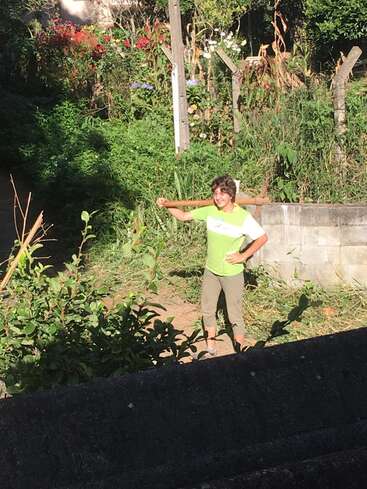 The image depicts a woman standing in a garden, holding a wooden tool behind her head, wearing a green shirt and khaki pants, with a concrete wall and lush vegetation in the background.