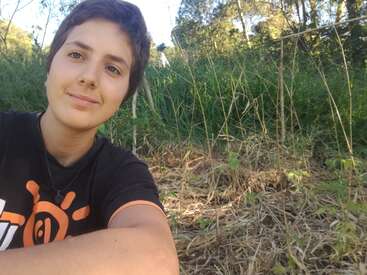 The image shows a young person with short hair, wearing a black t-shirt with an orange design, sitting in front of a grassy area with trees in the background.