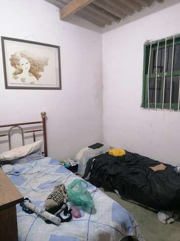 The image depicts a small, cluttered room with a bed, a window, and a painting on the wall, featuring a mix of personal items and clothing scattered about.