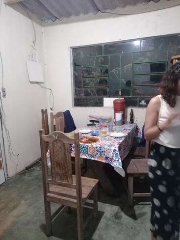 The image depicts a woman standing in a small, rustic room with a table and chairs, featuring a window and a white wall with a door on the left side.