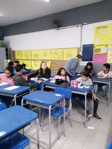 The image depicts a classroom with blue desks and chairs, featuring students engaged in an activity, accompanied by a teacher and colorful posters on the walls.
