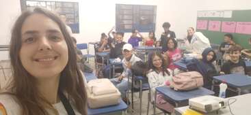 The image shows a group of students in a classroom, with a teacher standing at the front, surrounded by desks and chairs, conveying a sense of learning and education.