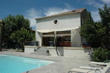 La imagen muestra una serena casa blanca con piscina y tumbonas, rodeada de árboles y un cielo azul con nubes difusas.