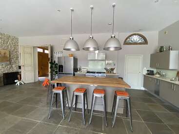 Esta moderna cocina cuenta con una isla de madera con cuatro taburetes metálicos, tres lámparas colgantes, electrodomésticos de acero inoxidable, paredes de piedra, armarios grises y elementos decorativos minimalistas.