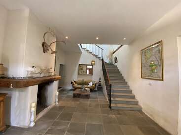 Esta imagen muestra un interior espacioso y rústico con suelo de piedra, escalera central, una acogedora zona de estar, chimenea, decoración con cuernos, cuadros en las paredes e iluminación tenue.