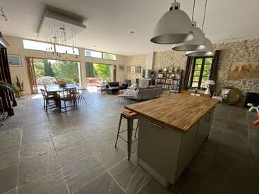 Esta espaciosa y moderna sala diáfana cuenta con una pared de piedra, grandes ventanas, una isla de cocina, zona de comedor, cómodos asientos, estanterías y mucha luz natural que entra por las ventanas.