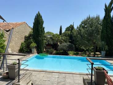 Una tranquila escena en un patio trasero muestra una piscina azul cristalina, exuberante vegetación, plantas en macetas, un edificio rústico de piedra y un cielo soleado y brillante.