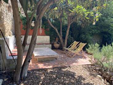 Un tranquilo patio exterior a la sombra de los árboles, con dos tumbonas, un banco de hormigón, una mesa baja, baldosas de terracota y rodeado de exuberante vegetación.