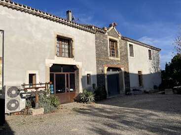 Esta imagen muestra una casa rústica de estilo mediterráneo con techo de tejas, detalles en piedra, grandes ventanas, plantas y un patio de grava bajo un cielo azul claro.