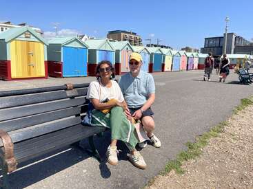 Una pareja se sienta en un banco junto a unas coloridas casetas de playa bajo un cielo azul claro. En el fondo, la gente pasea disfrutando de un día soleado.