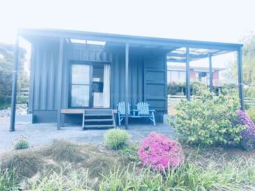 Ein modernes, kleines Haus, gebaut aus einem Schiffscontainer, mit einer überdachten Veranda, zwei blauen Stühlen, umgeben von üppigen Gartenpflanzen und bunten Blumen.