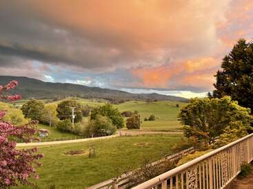 Eine wunderschöne Landschaft mit sanften grünen Hügeln, blühenden Bäumen, einer kurvenreichen Straße und dramatischen Wolken bei Sonnenuntergang, die die Szene in ein orange-graues Licht tauchen.