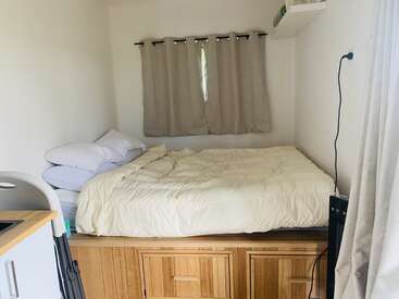 Ein gemütliches, minimalistisches Schlafzimmer mit einem erhöhten Holzbett, cremefarbenem Bettzeug, Kissen, beigen Vorhängen und einem kleinen Fenster. Helle Wände und eingebauter Stauraum schaffen Platz.