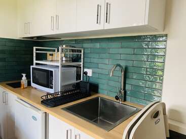 This image shows a compact kitchen corner with green tile backsplash, stainless steel sink, microwave, drying rack, soap dispenser, kettle, toaster, and white cabinetry.