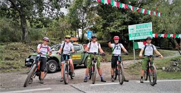 Das Bild zeigt eine Gruppe von Menschen auf Fahrrädern, mit einer Straße und Bäumen im Hintergrund. Die Szene spielt wahrscheinlich in einer natürlichen Umgebung im Freien, möglicherweise zu Freizeit- oder Wettkampfzwecken.