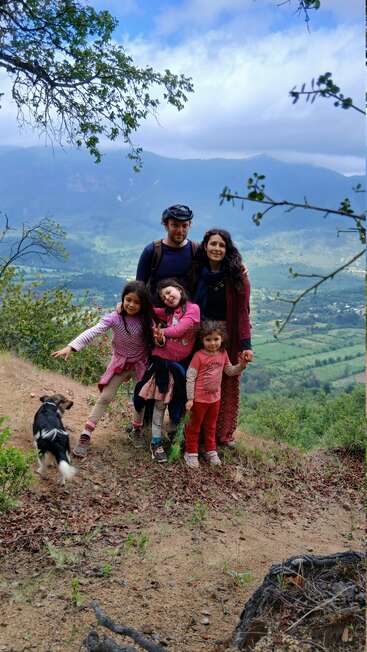Une famille de cinq personnes et un chien posent ensemble sur une colline pittoresque, entourés de verdure, de montagnes et d'un ciel nuageux spectaculaire, profitant de la nature et de la convivialité.