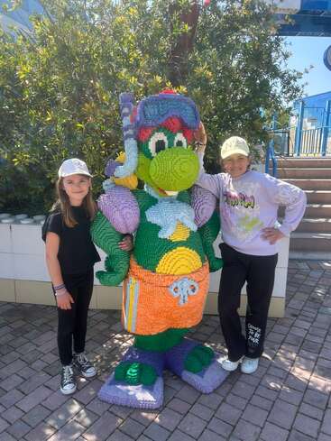 Two smiling girls pose with a large, colorful LEGO statue of a cartoon character wearing snorkeling gear and beach clothes at an outdoor amusement park or exhibit.