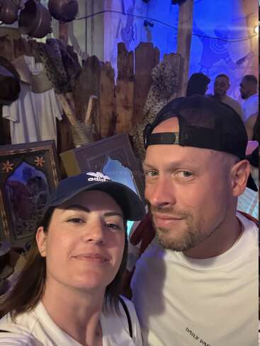 A woman in an Adidas cap and a man in a black cap pose together, smiling. Behind them, others gather in a warmly lit, rustic-themed room.