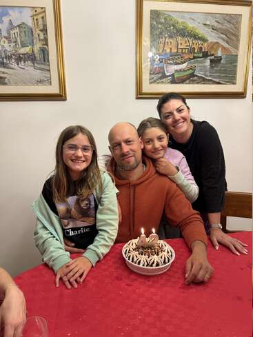 A joyful family celebrates a birthday together. The father, turning 42, sits at a table with a cake, surrounded by smiling loved ones in a cozy setting.