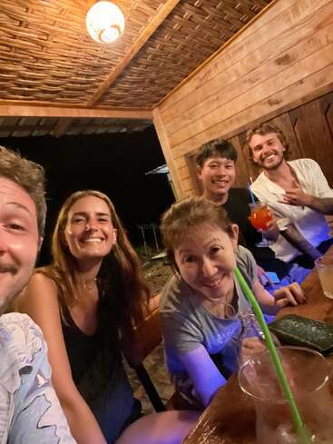 Das Bild zeigt eine Gruppe von fünf Personen, die an einem Tisch sitzen, lächeln und für ein Selfie posieren. Sie befinden sich in einem Holzgebäude mit einer gewebten Decke.