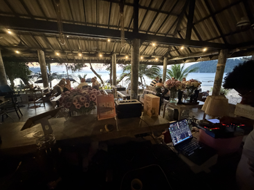 Ein gemütliches Restaurant am Strand bei Sonnenuntergang, voll mit Gästen. Lichterketten hängen von der Decke, tropische Pflanzen säumen den Bereich und ein DJ legt Musik auf.