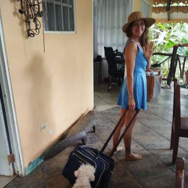 Une femme vêtue d'une robe bleue et d'un chapeau de soleil sourit et fait signe de la main, tirant une valise. Un petit chien blanc la suit à l'intérieur d'une maison tropicale confortable.