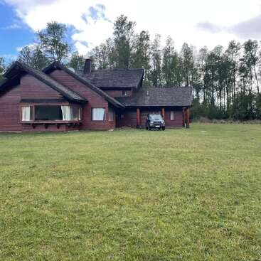 A rustic wooden house sits in a large grassy clearing, surrounded by tall trees. A black vehicle is parked under the covered porch. The sky is partly cloudy.