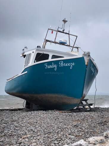 A blue and white fishing boat named “Fundy Breeze” rests on a rocky shore, with overcast sky and waves in the background, likely at low tide.
