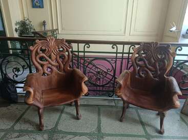Two intricately carved wooden armchairs sit side by side in a hallway, their organic, twisting designs evoking natural forms, backed by decorative wrought iron railing.