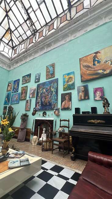 A spacious, artistic room with a glass ceiling, turquoise walls covered in colorful paintings, black piano, eclectic statues, checkered floor, vintage furniture, and a cozy, creative atmosphere.