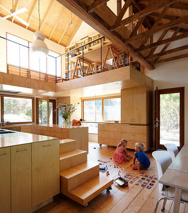The image depicts a spacious, open-plan room with a kitchen, dining area, and living space, featuring wooden floors and cabinets, and a lofted area with a railing.