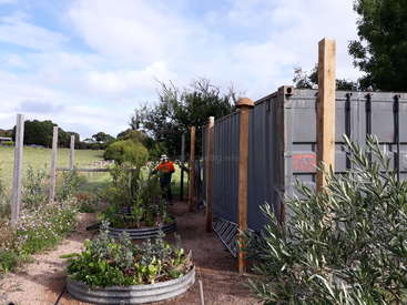 The image depicts a serene garden scene with a person tending to plants, surrounded by a fence and a shipping container, set against a picturesque rural backdrop.
