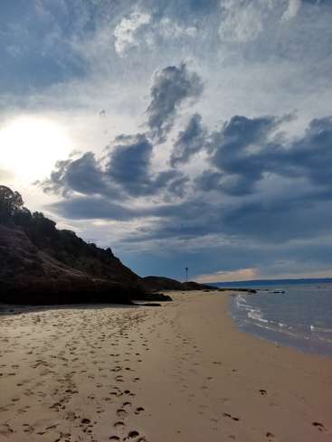 The image depicts a serene beach scene with a sandy shore, a rocky cliff, and a cloudy sky, featuring a body of water and a flagpole in the distance.