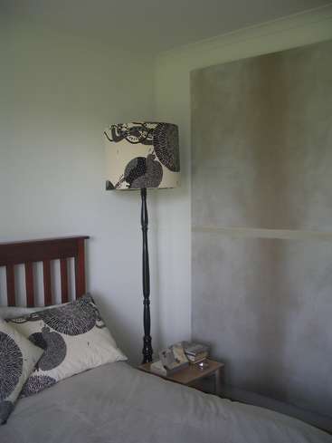The image depicts a serene bedroom corner featuring a bed with a dark wood headboard, a lamp with a black and white patterned shade, and a small nightstand with books and a candle.