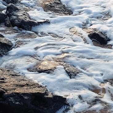 The image depicts a rocky shoreline with foamy water, featuring large dark rocks and a gentle flow of water, creating a serene and natural scene.