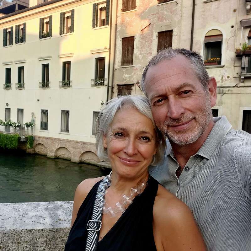 The image depicts a middle-aged couple posing for a photo in front of a canal and historic buildings in a European city, likely Venice, Italy.