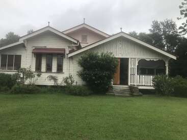 L'image représente une charmante maison blanche avec un porche, entourée d'une végétation luxuriante et d'une pelouse bien entretenue, sous un ciel couvert.