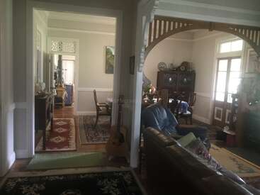 L'image représente une pièce spacieuse comprenant une salle à manger, un salon et un couloir, avec des murs blancs, des planchers en bois et divers meubles, dont un canapé et une guitare.