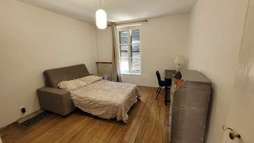 Cette chambre dispose d'un lit double, d'un sol en bois, de murs neutres, d'un petit bureau avec une chaise et une lampe, d'une commode métallique et d'une fenêtre avec des stores.