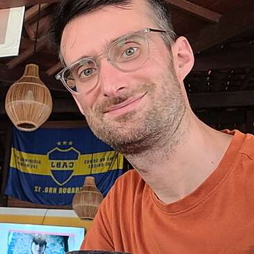 Un hombre con gafas y barba recortada sonríe a la cámara en un interior. Detrás de él hay lámparas colgantes, una bandera azul y amarilla y una pantalla de televisión.