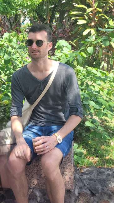 Un homme portant des lunettes de soleil, une chemise sombre et un short en jean est assis sur un banc en pierre, un verre à la main. Il est entouré de plantes tropicales verdoyantes.