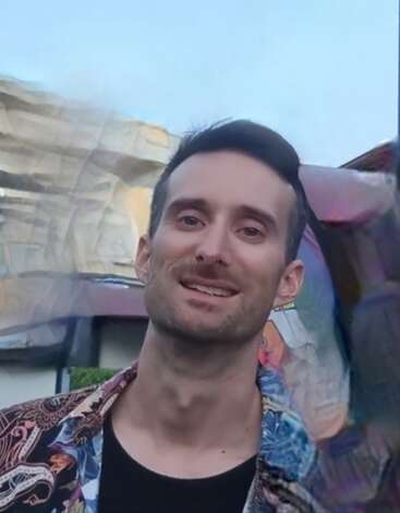 Um homem sorridente com cabelos escuros curtos está ao ar livre. Ele está vestindo uma camisa colorida. O fundo apresenta rochas e um efeito artístico desfocado. Céu azul acima.